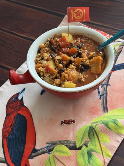 Rotes Thermoschüsselchen mit Griff, steht auf einem Set mit roten Vögeln. Im Schüsselchen ein scharfer karibischer Onepot.