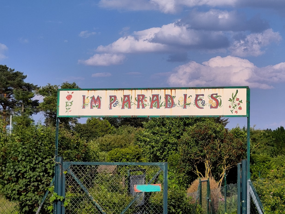 Foto: Über einem Gartentor aus Metall ist das Schild 'Im Paradies' angebracht. Die einzelnen Buchstaben sind mit Pflanzen- und Blumenoranmenten verschönert. Im Hintergrund Büsche und Bäume (ein Kleingartenkolonie). 