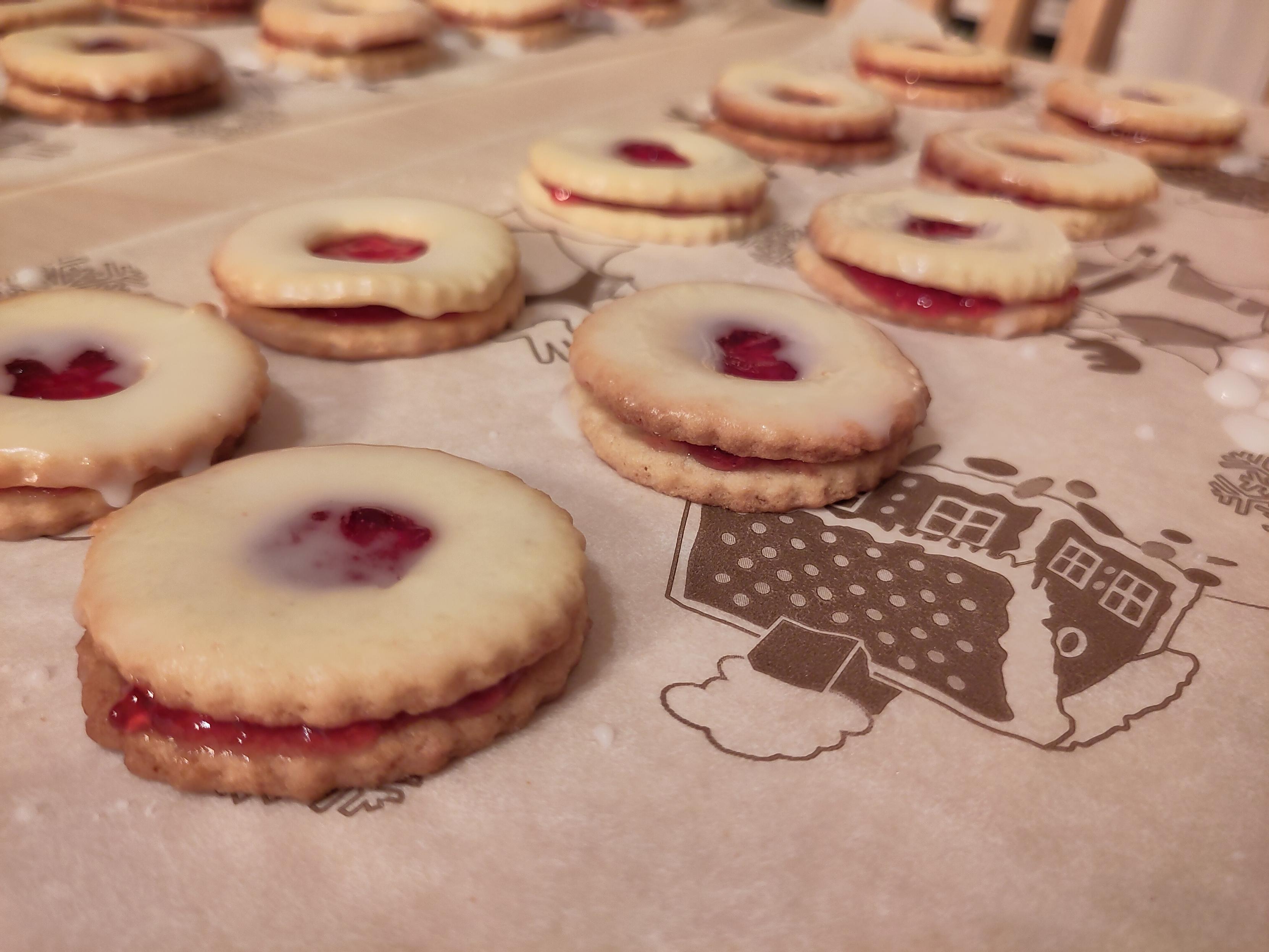 Auf einem Backpapier mit weihnachtlichem Motiv liegen Doppeldecker-Kekse, die in der Mitte mit Johannisbeerengelee gefüllt sind. Am Ende wurden sie mit Puderzuckerguss bestrichen.